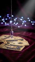 Purple crystal pendulum hanging over tarot cards on velvet fabric with bokeh lights creating mystical fortune telling atmosphere for spiritual guidance.