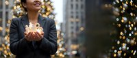 Young Asian businesswoman holding glowing lights in cupped hands during Christmas season in urban setting with festive decorations and bokeh background.