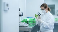 Female scientist in white lab coat and safety glasses examining green plant sample with magnifying glass in modern laboratory for botanical research and analysis.