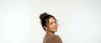 Young mixed-race woman with curly hair in bun smiling over shoulder wearing brown sweater on white background for lifestyle and fashion content.