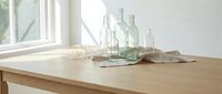 Minimalist wooden dining table with vintage glass bottles and linen cloth in bright modern kitchen interior with natural sunlight streaming through window.