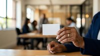 Professional holding blank white business card in modern office environment with colleagues meeting in background.