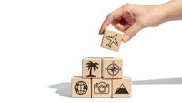 Hand placing wooden cube with airplane icon onto stack of travel-themed blocks on white background for tourism and vacation planning concepts.