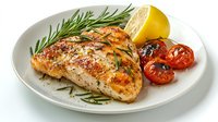 Grilled chicken breast seasoned with herbs and spices, served with fresh rosemary sprigs, roasted cherry tomatoes and lemon slice on white ceramic plate.