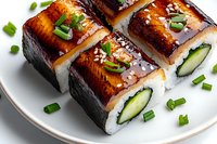 Grilled unagi sushi rolls with cucumber, rice and green onion garnish on white plate, glazed with sweet teriyaki sauce, close up view of Japanese cuisine.