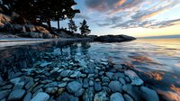 Dramatic winter shoreline with submerged rocks beneath frozen lake surface at sunset, reflecting vibrant orange clouds and blue sky in crystalline ice patterns.