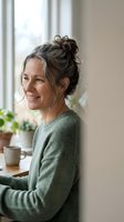 Smiling middle-aged woman with gray hair in messy bun wearing green sweater sitting by window in cozy home interior for lifestyle and wellness content.