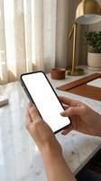 Hands holding smartphone with blank white screen mockup on marble desk with gold lamp and plants in bright home office.