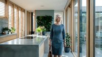 Mature woman with blonde hair wearing dark blue dress standing in modern kitchen with white island, wooden shutters, and potted plants for home design and lifestyle content.