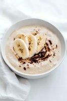 Creamy banana smoothie bowl topped with fresh banana slices and dark chocolate shavings on white background, healthy breakfast or dessert concept.