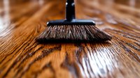 Natural bristle broom sweeping wooden floor surface, close-up view showing texture and grain pattern of hardwood planks in warm brown tones.