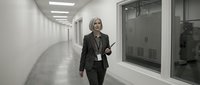 Middle-aged woman with blonde hair, Caucasian ethnicity, wearing business attire and ID badge, walking through modern corporate hallway with fluorescent lighting and glass offices.