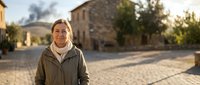 Middle-aged Caucasian woman smiling warmly in casual outdoor jacket standing on cobblestone street in charming European village during golden hour sunset.