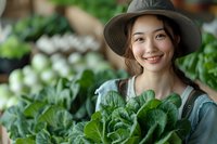 Young Asian woman in sun hat holding fresh organic leafy greens in greenhouse garden, representing sustainable farming and healthy lifestyle choices.