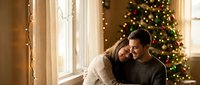 Young couple embracing by window with Christmas tree and warm lights creating cozy holiday atmosphere for seasonal romance and festive celebration.