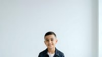 Young boy with dark hair and olive complexion, wearing a dark blue button-up shirt, smiling at camera against white background. Isolated portrait suitable for educational materials, family websites, and children's content.