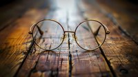 Round metal frame eyeglasses on rustic wooden surface with shallow depth of field. Vintage style optical accessories in warm lighting and bokeh background.