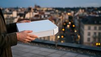 Woman holding blank white box on urban rooftop at dusk with city lights and buildings in background, suitable for product packaging mockups and delivery service marketing.