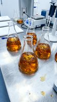 Laboratory flasks containing amber liquid samples arranged on white workbench with scientific equipment, notebooks, and instruments for chemical analysis and research testing.
