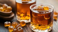 Amber-colored whiskey in crystal glass with ice cubes on rustic wooden table, golden hour lighting creates warm glow through liquid, selective focus.