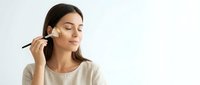 Young Caucasian woman applying foundation with makeup brush, eyes closed, natural beauty routine on white background.