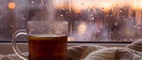 Steaming hot tea cup on windowsill during rainy evening with warm bokeh lights creating cozy atmospheric mood for relaxation and comfort at home.