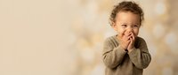 Adorable toddler boy with curly hair covering mouth with hands, giggling and playing peek-a-boo in cozy knit sweater against warm bokeh background.