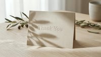 Elegant thank you card with embossed text on kraft paper, placed on a wooden table with olive branches and soft natural shadows.