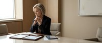 Mature blonde businesswoman in contemplative pose at modern office desk with documents and tablet for corporate planning and decision making.