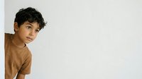 Young boy with dark curly hair and olive complexion wearing tan brown t-shirt, peeking around white wall corner with curious expression, isolated on white background.