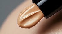 Liquid foundation makeup drop from applicator wand on beige skin surface showing creamy texture and smooth consistency for cosmetic and beauty industry.