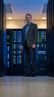 Confident middle-aged Caucasian businessman in suit standing in modern data center with glowing server racks for technology and corporate presentations.