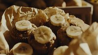 Freshly baked chocolate banana muffins topped with sliced bananas and nuts on brown parchment paper, warm morning breakfast setting in natural light.