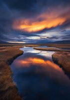 Dramatic sunset illuminates winding river through autumn grassland, with fiery orange clouds reflecting in calm water against moody blue sky and distant mountains.