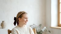 Young woman with blonde hair styled in braids with burgundy ribbon bow, wearing cream sweater, sitting indoors by window with natural light, smiling thoughtfully in minimalist home setting.