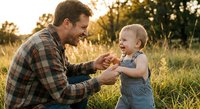 Father and toddler son playing together in golden hour sunlight outdoors in natural meadow setting during warm summer evening family bonding moment.