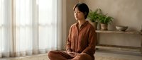 Young Asian woman in brown pajamas practicing mindfulness meditation sitting cross-legged on floor in bright modern bedroom with natural light and plants.