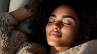 Young African American woman with curly hair sleeping peacefully in warm sunlight on soft bedding, expressing serenity and relaxation with gentle smile and closed eyes.