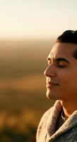 Middle-aged man with dark hair and olive skin tone in profile, wearing a beige sweater, gazing peacefully at golden hour sunset with soft warm lighting and blurred natural background.