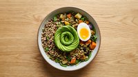 Healthy quinoa bowl with sliced avocado, soft-boiled egg, roasted vegetables and mixed greens on wooden table for nutritious meal planning.