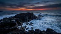 Dramatic rocky coastline with crashing waves during sunset, featuring dark jagged rocks and turbulent ocean waters under a moody sky. Ideal for travel and nature projects.