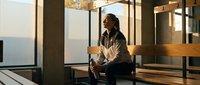 Young African American businesswoman sitting on modern office stairs during golden hour, holding coffee cup and looking thoughtfully out window in contemporary workplace.