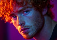 Young caucasian male with ginger curly hair and blue eyes in dramatic neon lighting. Close up portrait showing intense expression against purple and orange background.