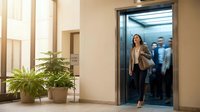 Young professional woman in beige blazer stepping out of modern office elevator in contemporary business building lobby with plants and natural lighting.