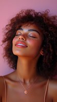 Young African American woman with curly hair and glowing skin smiling with eyes closed against pink background, wearing delicate necklace and natural makeup with freckles.