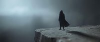 Silhouette of person in dark coat standing on cliff edge overlooking misty valley in moody atmospheric landscape with dramatic stormy sky and fog.