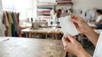 Hands holding blank white card in creative studio workspace with fabric swatches, design materials, and colleagues working in background for branding and business concepts.