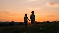 Two children holding hands standing in an open field at sunset, silhouetted against a warm golden sky with clouds, outdoor leisure and childhood bonding moment.