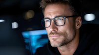 Professional Caucasian businessman with glasses working late at night in modern office with computer screen glow reflecting on face for corporate projects.
