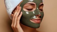 Young woman with green clay mask on face and white towel on head enjoying spa skincare treatment. Close up beauty portrait showing glowing skin and manicured nails.